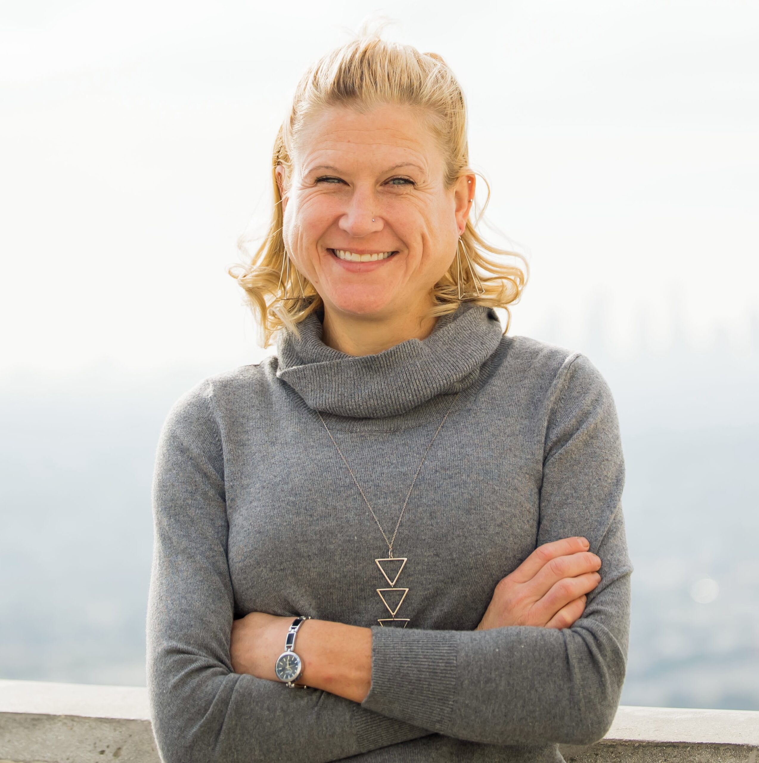 A white middle-aged woman with blonde hair and blue eyes smiles outside in front of a white building and green hedges.